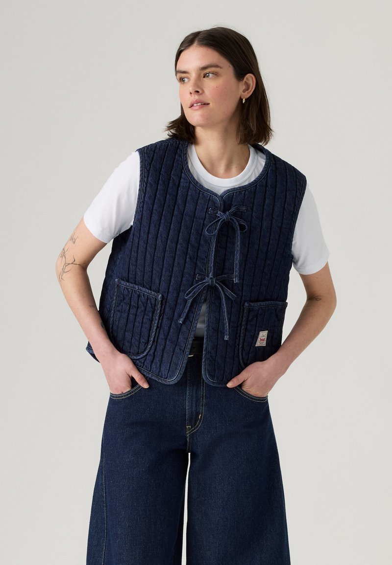 Chaleco acolchado azul oscuro con escote redondeado, lazos frontales y dos bolsillos en la parte delantera, usado sobre una camiseta blanca y combinado con jeans anchos oscuros.