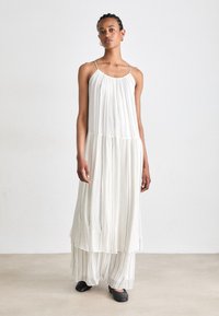 Weißes, plissiertes Maxikleid mit dünnen Trägern und geschichtetem Design. Verfügt über vertikale schwarze Streifen und fließt bis zu den Knöcheln. Kombiniert mit schwarzen Schuhen.