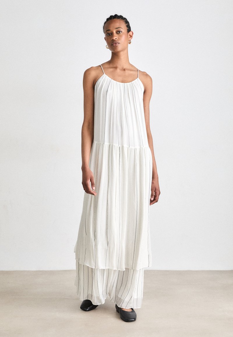 Weißes, plissiertes Maxikleid mit dünnen Trägern und geschichtetem Design. Verfügt über vertikale schwarze Streifen und fließt bis zu den Knöcheln. Kombiniert mit schwarzen Schuhen.