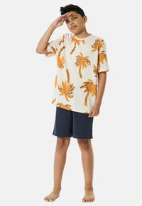 Camicetta a maniche corte color crema con stampa di palme arancioni, abbinata a pantaloni corti blu navy, indossata da un bambino a piedi nudi che saluta.