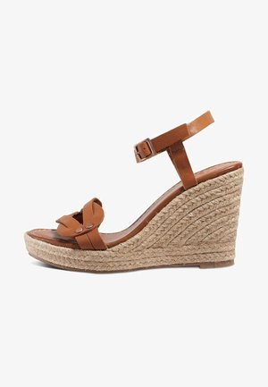 Brązowy skórzany sandał na koturnie z paskiem wokół kostki i plecioną podeszwą z juty, odkrytym przodem i ozdobnym supłem na przednim pasku.