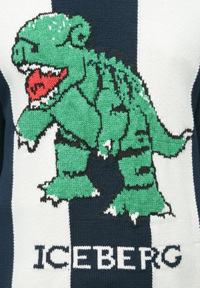 Πράσινος καρτουνίστικος δεινόσαυρος με κόκκινο στόμα και λευκά δόντια πάνω σε πλεκτό ύφασμα με ναυτικό μπλε και λευκές ρίγες, με το κείμενο «ICEBERG» από κάτω.
