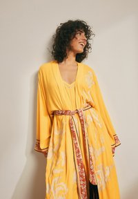Geltonas kimono stiliaus chalatas su gėlių raštais, plačiomis rankovėmis ir kontrastinga raudona bei balta juosta juosmeniui, papuošta kutais.
