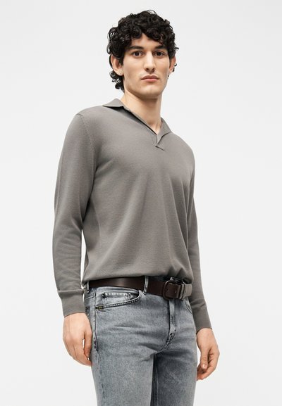 Νέος άνδρας με σκούρα σγουρά μαλλιά που φοράει γκρι μακρυμάνικο πόλο πουκάμισο μέσα σε ανοιχτόχρωμα γκρι τζιν με καφέ δερμάτινη ζώνη.