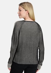 Kobieta z blond włosami, ubrana w czarno-białą, pionowo prążkowaną długosleevową bluzę, stojąca bokiem z delikatnym uśmiechem.