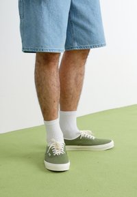 Gröna canvas sneakers med vita sulor, tillsammans med vita ankelsockor och ljusblå denimshorts, på ett grönt golv.