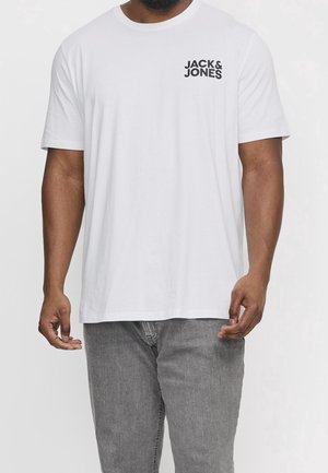 Homme portant un T-shirt blanc à manches courtes avec texte noir "JACK & JONES" sur la poitrine et un jean gris, debout devant un fond uni.