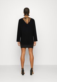 Vestido preto de mangas longas com detalhes em renda nas costas, apresentando um recorte em forma de gota. Textura suave e comprimento acima do joelho. Saltos visíveis.