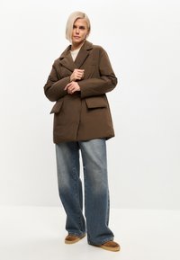 Chaqueta acolchada marrón con cuello de solapa, dos bolsillos frontales y un corte relajado. Combinada con jeans de denim azul de pierna ancha y zapatos marrones sin cordones.