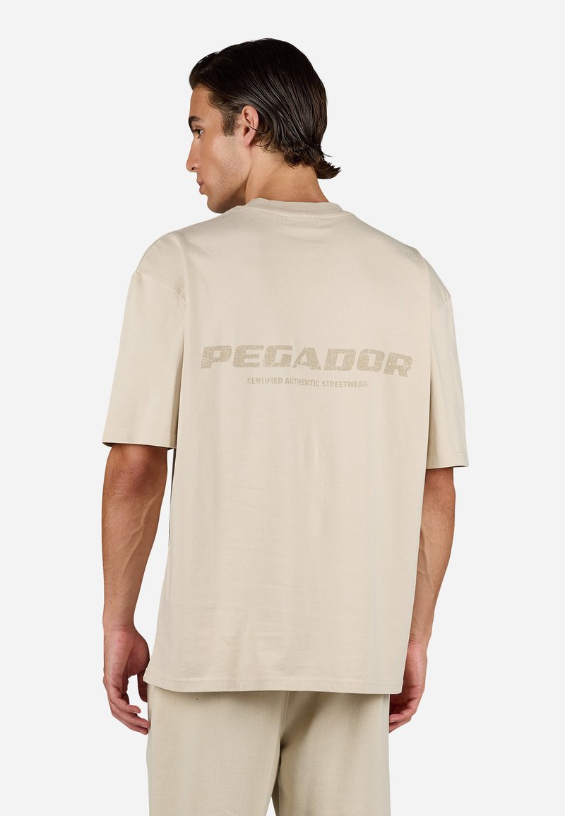 Beige oversized T-skjorte laget av myk stoff, med en stor, teksturert "PEGADOR" logo og "CERTIFIED AUTHENTIC STREETWEAR" trykket på baksiden.