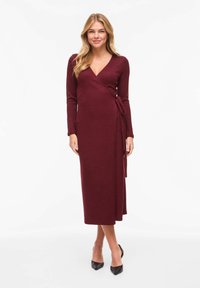 Robe portefeuille bordeaux mi-longue à manches longues, avec une ceinture à nouer à la taille. Fabriquée en tissu doux et texturé. Escarpins noirs à bouts pointus.