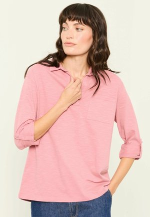 Woman with dark wavy hair wearing a pink long-sleeve shirt with rolled cuffs and blue jeans, looking to her left with one hand near her collar.