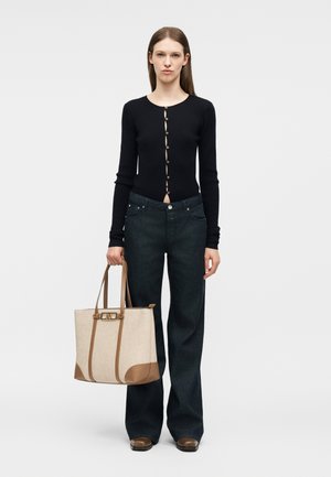 Γυναίκα που φοράει μαύρη ζακέτα με κουμπιά, φαρδιά σκουρόχρωμα τζιν, καφέ παπούτσια, κρατώντας μπεζ και καφέ τσάντα tote, στέκεται μπροστά σε λευκό φόντο.