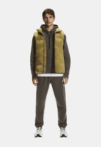 Junger Mann in braunem Hoodie, olivgrünem Steppwesten, passenden Jogginghosen und weißen Turnschuhen, steht vor weißem Hintergrund.