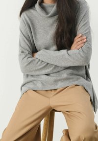 Persona seduta su uno sgabello di legno con le braccia incrociate, che indossa un maglione grigio e pantaloni beige su uno sfondo semplice.