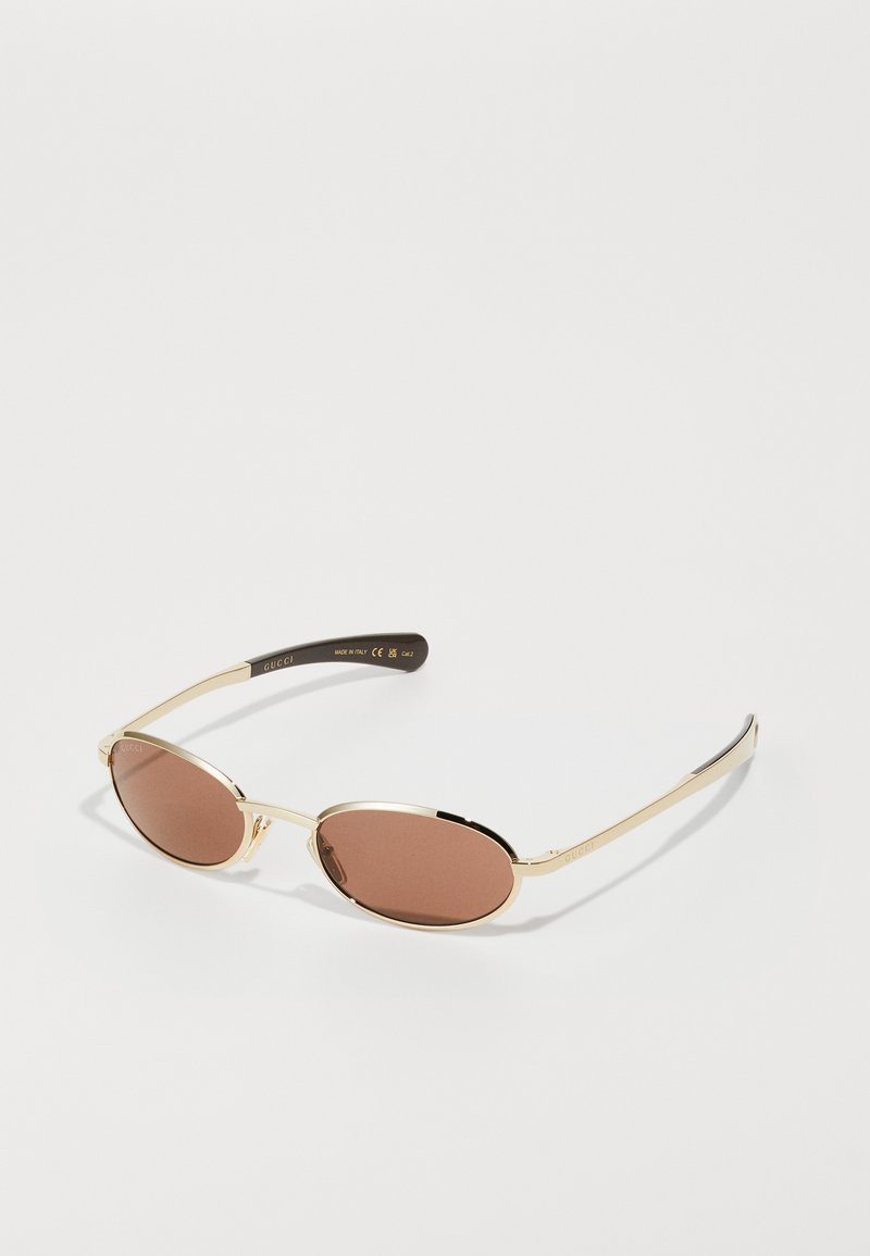 Zlaté oválné sluneční brýle s hnědými tónovanými skly. Úzký kovový rám s hladkým povrchem, který má černé akcenty na spánkách a gravírované logo.