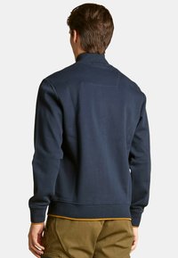 Sudadera azul marino con cuello alto, puños y dobladillo acanalados. Presenta un sutil detalle de costura en la parte posterior y un borde en mostaza contrastante en la parte inferior.