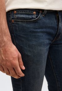 Κοντινή εικόνα του χεριού ενός άνδρα που ακουμπάει δίπλα σε σκούρο μπλε τζιν με εμφανείς ραφές και χαλκομανίες.