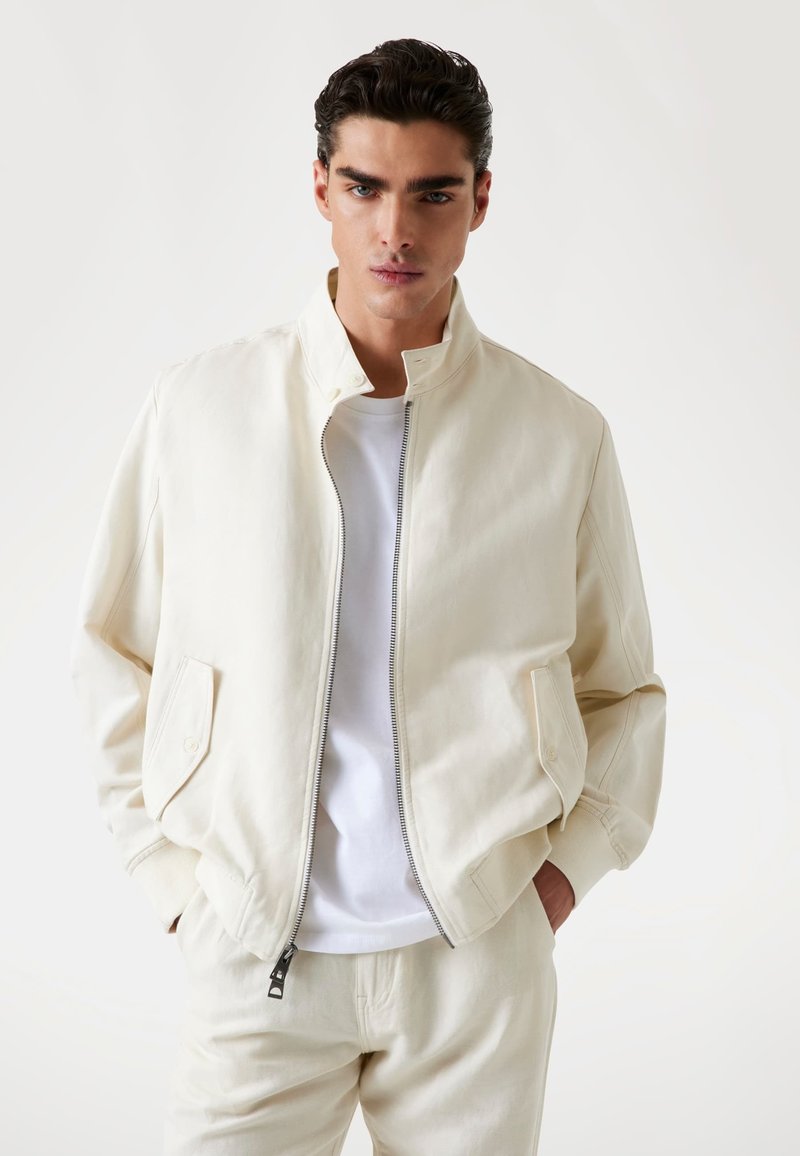 Light beige bomber jacket made of cotton, featuring a high collar, front zip, and side pockets. Worn over a white t-shirt.