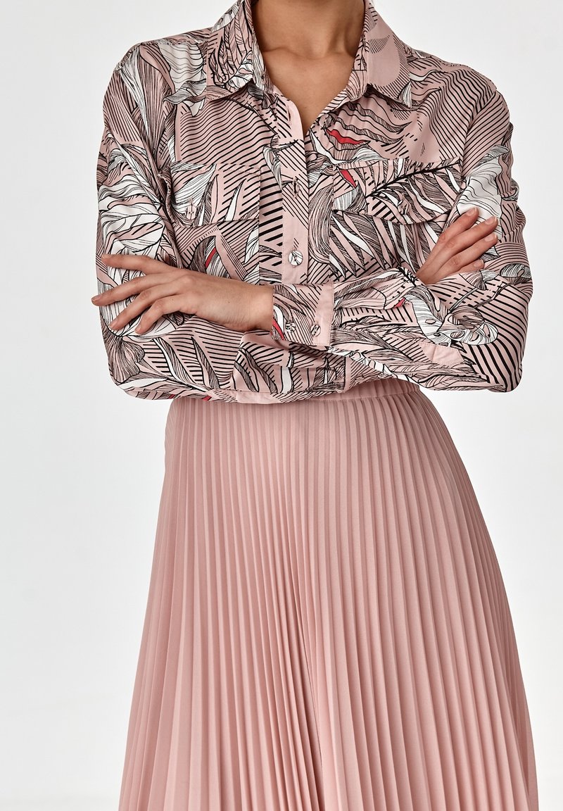 Blusa a maniche lunghe di un rosa chiaro con motivi tropicali di foglie nere e bianche, abbinata a una gonna plissettata di un rosa pallido, che mette in risalto movimento e texture.