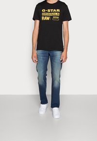 Černé krátké tričko s žlutým grafickým textem, spárované se světle modrými džínami s vybledlým designem a bílými teniskami.