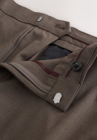 Pantalon marron avec fermeture éclair, bouton et fermoir en métal, montrant des coutures détaillées et une doublure intérieure avec bordure bordeaux.