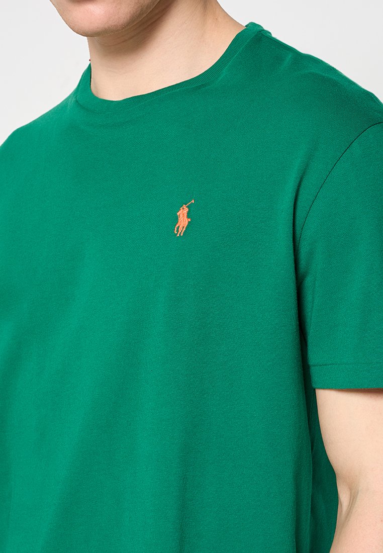 Mężczyzna ubrany w zielony t-shirt z krótkim rękawem z pomarańczowym haftowanym logo gracza polo na piersi.