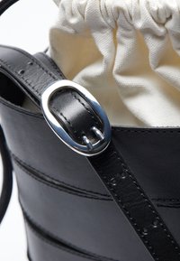 Mustasta nahkabägistä, jossa on kerrottu vaaleanruskea kangasvuori, ja jossa on hopeinen soikea metalliosuus teksturoidussa nahkahihnassa.