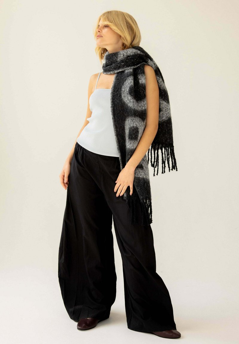 Écharpe noire et grise à motifs avec franges, fabriquée en tissu doux et texturé, portée sur un haut léger et un pantalon noir ample.