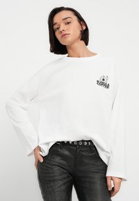 Νεαρό άτομο με κοντά σκούρα μαλλιά φοράει λευκό πουκάμισο με μακριά μανίκια με το λογότυπο «Bagels & Coffee» και μαύρο υφασμάτινο παντελόνι με ζώνη με τρουκς.