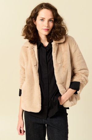 Beige nepbontjas met een kraag, zijzakken en een ontspannen pasvorm, gecombineerd met een zwart shirt en donkere spijkerbroek. Zachte textuur.