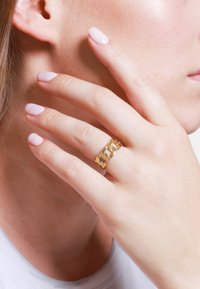 Goldene Kettenring an einem Finger, mit glattem, poliertem Metall und einem verbundenen Muster; Hautfarbe und hellrosa Nägel sichtbar.