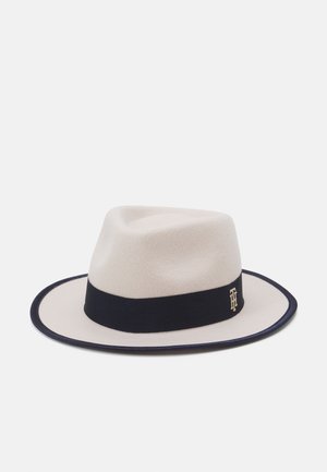Sombrero fedora color crema con banda y ribete negro, con un pequeño emblema dorado en la banda, sobre un fondo blanco liso.