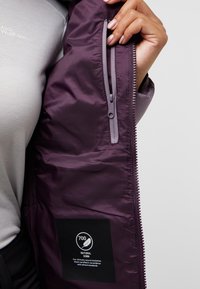 Giacca isolante viola con tasca con zip, superficie liscia e un'etichetta nera che indica un'imbottitura in piuma d'anatra naturale con potenza di riempimento 700.