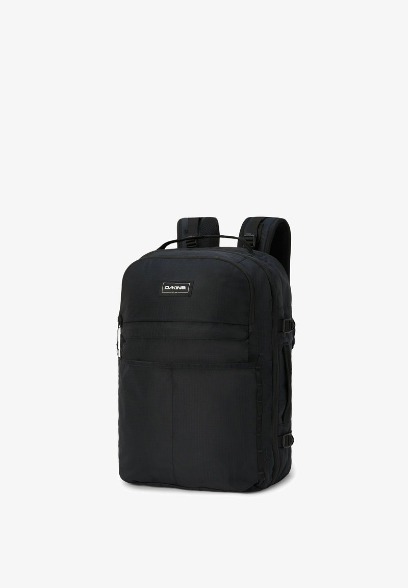 Svart Dakine-ryggsäck med flera zip-fickor och vadderade axelremmar, stående upprätt mot en vit bakgrund.