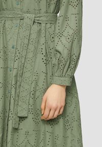 Abito lungo verde realizzato in tessuto texturizzato, con dettagli ad occhio di pernice, vita con cinturino, maniche a sbuffo lunghe e chiusure a bottone.
