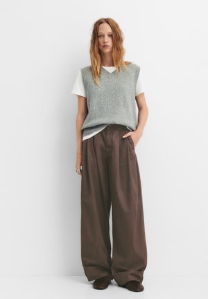 Un gilet lavorato a maglia grigio chiaro sopra una maglietta bianca, abbinato a pantaloni marroni larghi e scarpe marroni. Il modello è in piedi con le mani nelle tasche.