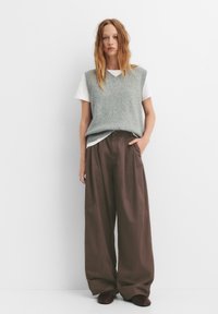 Lichtgrijze gebreide vest over een wit T-shirt, gecombineerd met losse bruine broek en bruine schoenen. Model staat met handen in de zakken.