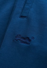 Tissu bleu foncé avec un logo brodé indiquant "SuperDry" et un petit détail de poche sur le côté supérieur gauche.