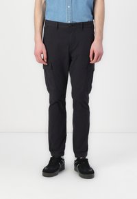 Cargo-byxor i mörkgrå bomullsblandning med fram- och sidofickor, tapered design och knappstängning. Bärs med svarta sneakers.
