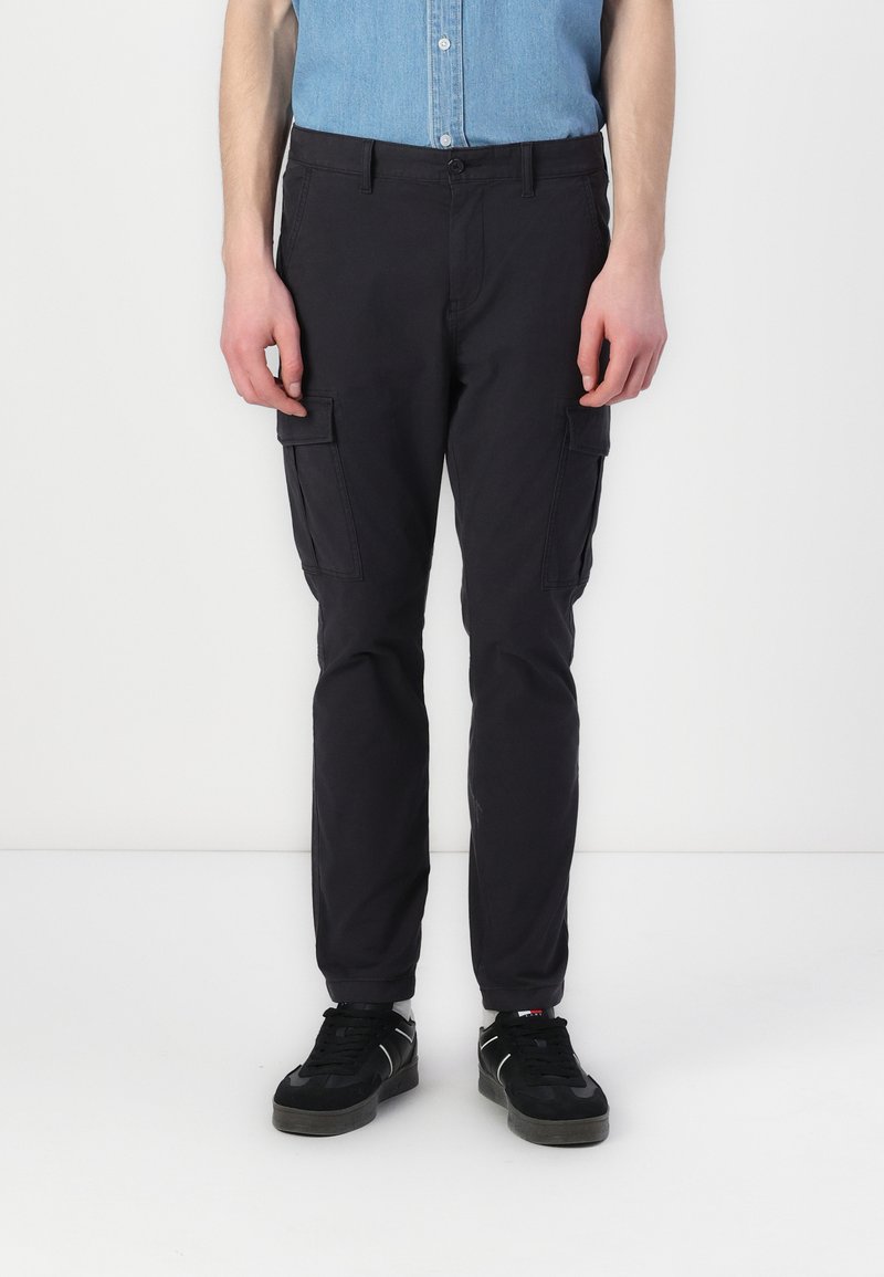 Cargo-byxor i mörkgrå bomullsblandning med fram- och sidofickor, tapered design och knappstängning. Bärs med svarta sneakers.