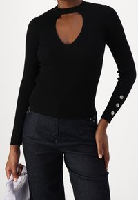 Haut noir à manches longues avec côtes, présentant une encolure à découpes et des boutons argentés sur les manches, porté avec un pantalon en denim foncé et un sac à main clair.