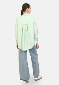 Camisa de manga larga verde claro con pliegue en la espalda y detalle de lazo. La tela es suave, con un dobladillo curvado, combinada con pantalones de mezclilla de pierna ancha.