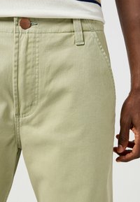 Pantaloni in cotone verde chiaro con tasca anteriore, cuciture visibili e pulsante con logo marrone. La texture appare liscia e morbida.