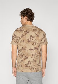 T-shirt à manches courtes beige avec motif camouflage marron, col rond, texture douce et coupe décontractée.