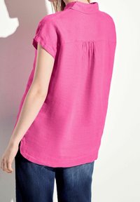 Blusa rosa a maniche corte con colletto, caratterizzata da una piega sul retro e un taglio rilassato, abbinata a jeans blu scuro. Tessuto morbido e texture.