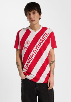 Jong persoon met kort donker haar die een rood-wit gestreept sportshirt draagt met de tekst "DANISH DYNAMITE" en zwarte broek.