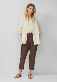 Crème button-down overhemd over een beige top, gecombineerd met bruine tapered broek en bruine sandaaltjes met dierenprint. Casual, relaxed fit.