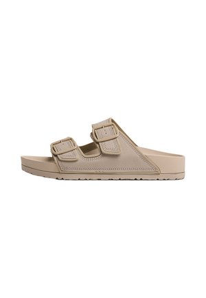 Sandalia beige tipo slide con dos tiras ajustables con hebilla y una suela texturizada para agarre, mostrada de lado.