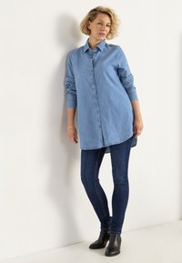 Helles blaugraues Jeanshemd mit durchgehender Knopfleiste, langen Ärmeln und asymmetrischem Saum, kombiniert mit dunkelblauben Skinny Jeans und schwarzen Stiefeletten.